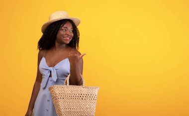 Saman çantalı ve şapkalı Afro-Amerikalı kadın turuncu stüdyo arka planında boş bir alanı işaret ediyor, afiş tasarımı. Mutlu müşteri, mevsimlik indirim, özel teklif konsepti