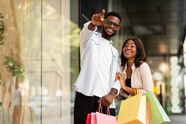 Müşteri ve Tüketim Konsepti. Gülümseyen Afro-Amerikan çift alışveriş merkezinin önünden birlikte yürüyüp dışarıyı gösteriyorlar. Mevsimlik Satış ve İndirim, Satın Alım ve Perakende