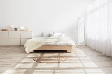 Ev tasarımı, minimalizm ve sabah içiyle ilgili fikirler. Yastıklı yatak, yumuşak battaniye, halı, ayna, ahşap zeminde mobilya. Beyaz duvar, yatak odasında perdeleri olan büyük pencere, hiç kimse, boş alan