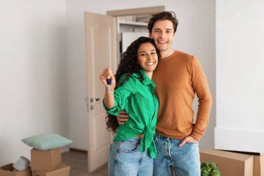 Neşeli çift yeni dairelerinde birlikte duruyorlar, anahtarlarını tutuyorlar ve taşındıktan sonra gülümsüyorlar. Taşınan kutuların arasında kucaklaşıp ev sahipliği yolculuklarının başlangıcını kutluyorlar..