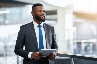 Takım elbiseli bir adam elinde tabletle modern bir ofis binasında duruyor ve gülümsüyor. Ayarlar, geniş pencereli ve doğal ışığın içeri girdiği parlak, açık bir alan gösteriyor..