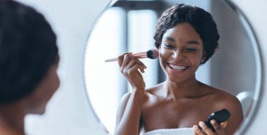 Bir kadın evinde yuvarlak aynanın önünde fırça kullanarak makyaj yaparken gülümsüyor. Mutlu bir ifade sergiliyor ve sabahları güzellik rutininden zevk alıyor..