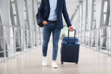 El çantalı tanınmayan bir adam havaalanı terminalinde yürürken günlük kıyafetleriyle uçağa bagajla binen, iş seyahatine çıkmaya hazır genç bir adamın kırpılmış fotoğrafı.