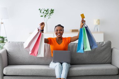 Başarılı internet alışverişi. Poşet ve kredi kartı tutan pozitif siyahi kadın evdeki kanepede otururken online sipariş vermek için laptop kullanıyor. E-ticaret kavramı