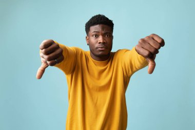 Üzgün Afro-Amerikan milenyumu sarı hırkalı adam başparmağını kaldırıyor ve mavi stüdyo arka planıyla ilgili hayal kırıklığını dile getiriyor, bir şeyi beğenmiyor, kopyalama alanı. İnsan jestleri kavramı
