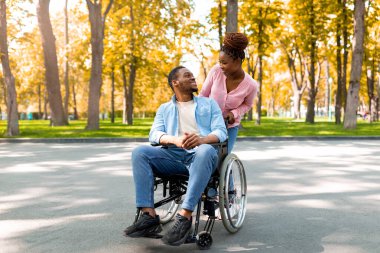 Sevecen zenci kadın ve tekerlekli sandalyedeki engelli erkek arkadaşı sonbaharda şehir parkında yürürken birbirlerine bakıyorlar. Mutlu engelli çift, doğanın tadını çıkarıyor.