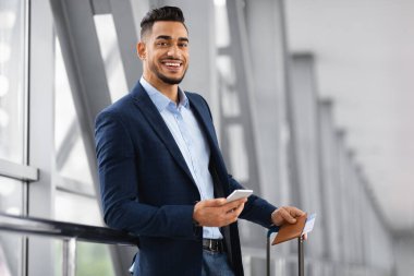 Havalimanı terminalinde uçuşu bekleyen yakışıklı Orta Doğulu iş adamının portresi, bagajı ve akıllı telefonu olan genç bir Arap, iş gezisine hazır, kopyalama alanı.