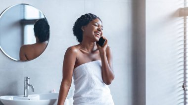 Bir kadın banyoda havluya sarılmış, telefonla konuşurken gülümsüyor. Doğal ışık pencereden girer ve parlak bir atmosfer yaratır..