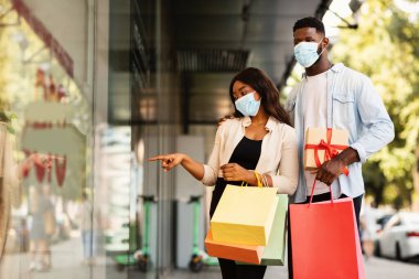 Hadi bunu alalım. Tıbbi maske takan Afro-Amerikan kadın portresi dükkan camını işaret ediyor, alışveriş merkezinin yakınında kocasıyla birlikte çantalarla yürüyor. Mevsimlik Satış ve İndirim, Özel Teklif