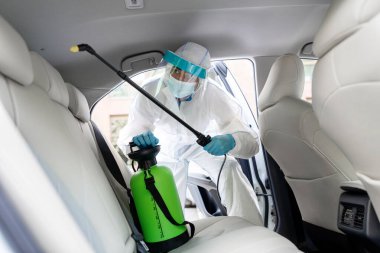 Özel koruyucu ekipman PPE kullanan bir temizlikçi arabanın içindeki kimyasal alkol spreyini kullanarak CV19 'u dezenfekte ediyor. Tehlikeli madde giysisi giyen adam otomatiği dezenfekte eder ve temizler.