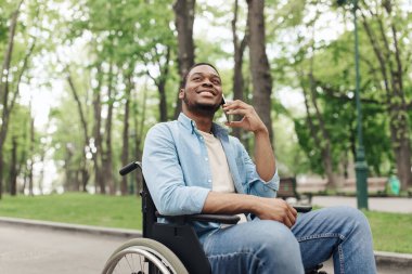 Tekerlekli sandalyedeki olumlu genç adam şehir parkında akıllı telefondan konuşuyor, kopyalama alanı. Milenyum Afrikalı Amerikalı adam cep telefonuyla dışarıda konuşuyor.
