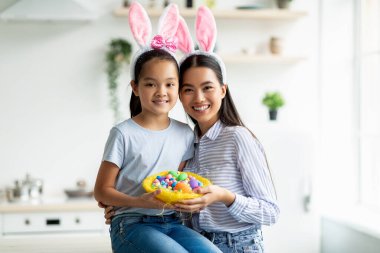 Mutlu Paskalyalar. Güzel Asyalı kız ve kadın tavşan kulağı takıyor ve ellerinde boya dolu yumurta sepeti var. Mutfakta otururken kameraya sarılıp gülümsüyorlar.