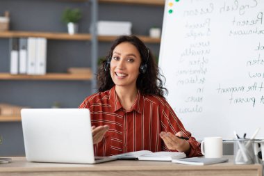 Uzaktan Öğrenme Konsepti. Gülümseyen öğretmenin portresi kameraya kablosuz kulaklıklar takarak konuşuyor ve ekranın yanında bilgisayar başında oturuyor. Eğitici materyalleri açıklayan kadın