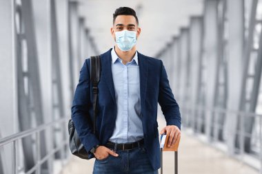Coronovirus Seyahatleri. Tıbbi maskeli genç bir adam havaalanı binasında bavuluyla duruyor. Salgın sırasında seyahat eden orta doğulu bir adam. Pasaportu elinde biletlerle kameraya bakıyor.