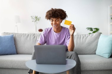 Uzaktan alışveriş konsepti. Bilgisayarı ve kredi kartıyla evden mal sipariş eden pozitif siyah bir genç. Neşeli Afrikalı Amerikalı ergen oturma odasında internetten bir şeyler alıyor.