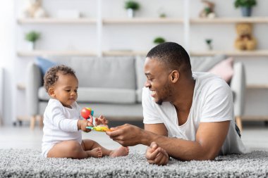 Bebeklerle Gelişim Faaliyetleri. Genç siyahi babanın evde bebekle oynadığı portresi, mutlu Afrikalı Amerikalı adam sevimli bebeğine oyuncak veriyor ve gülümsüyor.
