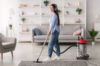 Evini süpürürken, elektrikli süpürge kullanarak kablosuz kulaklıkla müzik dinleyen neşeli genç bir kadın. Mutlu milenyum hanımları şarkı söylüyor, ev işlerinden zevk alıyor.