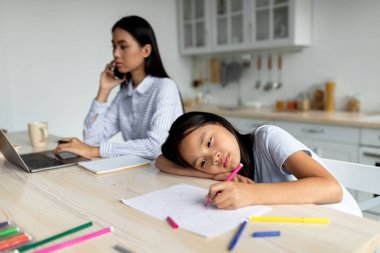 Can sıkıntısı konsepti. Evde uzaktan çalışan ve kızını görmezden gelen meşgul bir anne. Sıkılmış ve yalnız hisseden çocuk, kağıda çizim yapıyor. Asyalı anne ve kızı mutfakta oturuyor.