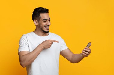 Happy bearded millennial arabic guy pointing at mobile phone screen and smiling, having video chat with friends, using mobile app for telecommunication, yellow studio background, copy space