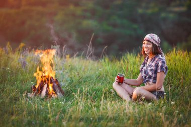 Kız şenlik ateşi yakın. Seyahat, turizm, kamp - genç ince sportif kadın turist esmer çadır ateşin yanında oturan güzel doğa manzaraya. Kız onun elinde kupa içki ile.