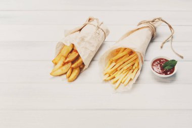 Patates dilimleri ve patates kızartması kahverengi ambalaj kağıdına sarılmış. Fast food beyaz eski şık ahşap götürmek. Domates soslu kızarmış patates. Cips, patates dilimleri. Üst görünüm, copyspace ile düz yatıyordu