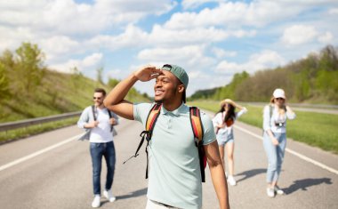 Genç siyahi adam arkadaşlarıyla yolda yürüyor, mesafeye bakıyor, turist gezisine çıkıyor. Otostop, yaz yolculuğu macera, arkadaşlık ve otostop konsepti