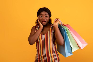 Excited black female with shopping bags touching her face in surprise, cannot believe huge sale on orange studio background. Pretty African American woman buying on discount