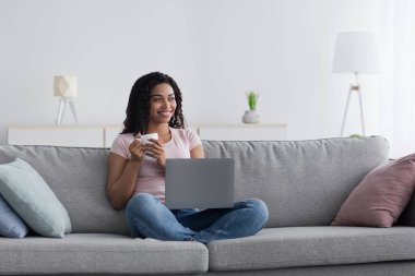 Modern cihazlarla evde dinlen, evde çalış ve kahve molası ver. Bin yıllık mutlu Afro-Amerikan kadın dizüstü bilgisayarı ve bir fincan sıcak içeceği olan oturma odasındaki boşluğa bakıyor.