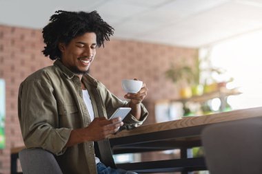 Kahvesini yudumlayan ve kafede otururken akıllı telefon kullanan neşeli siyahi adam cep telefonuyla internete göz atan ya da internetten Morning News okuyan Afrikalı genç bir Amerikalı.