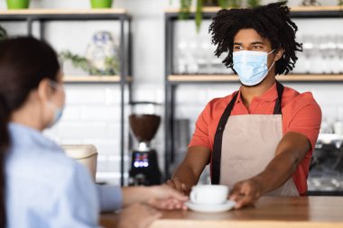 Gülümseyen Afro-Amerikan kıvırcık erkek garson koruyucu maske ve önlükle müşterilere kahve tezgahında taze kahve veriyor. İyi hizmet, iş, yeni normal ve çalışma süresi covid-19