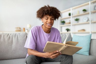 Açık kitabı olan, rahat koltukta oturan, ders çalışan ya da ev ödevi yapan pozitif bir genç. Oturma odasında kitap okuyan Afro-Amerikan ergenlerin portresi.