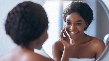 Banyoda aynanın önünde duran ve cilt bakımı yapan biri. Ayar, içinden doğal ışık geçen iyi ışıklandırılmış bir alan gösteriyor..