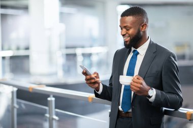 Resmi kıyafetli bir adam ofis binasında telefonuna bakıyor. Bir elinde kahve fincanı tutuyor ve gülümsüyor. Çevre parlak ve modern, bu da yoğun bir iş günü olduğunu gösteriyor..