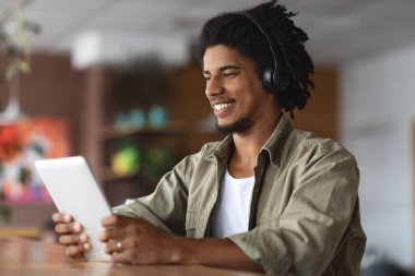 Kafede dijital tablet kullanarak kablosuz kulaklık takan mutlu siyah adam, modern kahve dükkanında masada otururken gülümseyen Afrikalı Amerikalı genç adam,