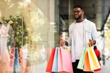 Tüketim kavramı. Afro-Amerikan bir adamın alışveriş merkezinin vitrinindeki mankenlere bakmasına sevindim. Elinde alışveriş poşetleri tutan gülümseyen siyah adam, fotokopi alanı, bulanık arka plan. Perakende ve Satın Al