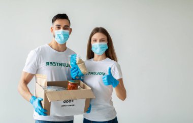 İki mutlu genç gönüllü, yiyecek bağış kutusunu tutarken tıbbi maske takıyor. Bir gönüllü onay veriyor, diğeri de tahıl paketini sunuyor. Işık duvarına karşı dururlar..