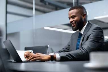 Adamın biri parlak bir ofiste, bilgisayarına odaklanmış bir masada oturuyor. Gülümserken kağıda notlar yazıyor. Ayarlar cam duvarlı çağdaş bir çalışma alanı gösteriyor.