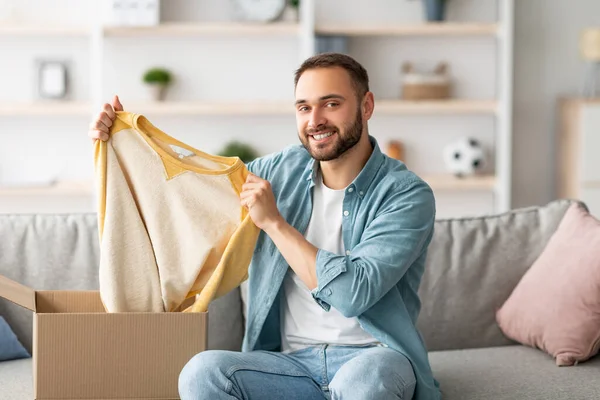 Karton kutudan yeni bir kazak çıkaran tatmin olmuş erkek müşteri, evdeki teslimat servisinden memnun. Millennial elemanı karton paketleri açıyor, internetten sipariş alıyor, kapalı yerlerde.