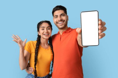 Bir çift canlı mavi bir arkaplan önünde birbirlerine yakın durur, gururla bir akıllı telefonu sergilerken gülümserler. Onların mutlu ifadeleri, özel bir anda heyecan ve neşe taşır..