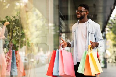 Satış ve indirim, perakende satış, alım. Gülümseyen Afrikalı Amerikalı adamın portresi alışveriş merkezinin vitrinindeki moda kıyafetlerine bakıyor, alışveriş merkezinin yakınında yürüyor, elinde hediyelerle alışveriş torbaları tutuyor.