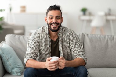 Genç gülümseyen Arap adam evdeki koltukta sıcak aromatik kahve içiyor, fotokopi çekiyor. Ilık içeceklerle yakışıklı Doğulu adam mola veriyor, oturma odasında tembellik ediyor.