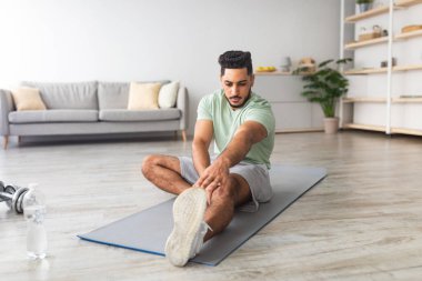 Paspasın üzerinde oturan genç bir Arap, evde bacağını esnetiyor, uzun bir portre. Milenyum Orta Doğulu adam esnek karantina sırasında oturma odasında antrenman yapıyor.