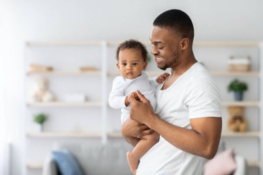 Mutlu Afrika kökenli Amerikalı baba, evde bebeğiyle poz veriyor, genç siyah babaya bakıyor, kucağında küçük şirin bir çocuğu kucağında kucağında taşıyor ve sıcak oturma odasında duruyor, boşluğu kopyalıyor.