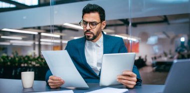 Bir işadamı modern bir ofiste masa başında oturuyor, bir elinde tablete bakarken diğer elinde belgeleri inceliyor. Çalışma alanı bitkiler ve parlak ışıklandırma içerir.