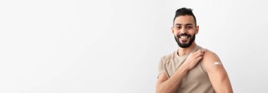 Coronavirus aşısı hayat kurtarıyor. Neşeli bir Arap adam koluna yara bandı takıyor. Aşı aşısı yaptıktan sonra gülümsüyor, beyaz stüdyo arka planında korunduğunu hissediyor.