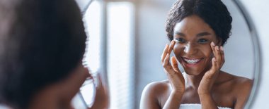 Parlak bir banyoda aynaya bakan bir kadın. Yüzüne cilt bakımı yaparken gülümsüyor. Güneş ışığı bir pencereden odaya girer..