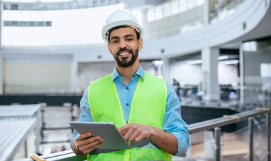 A construction worker stands indoors at a building site. He wears a safety vest and helmet while looking at a tablet. The area is busy with activity and construction materials around him.