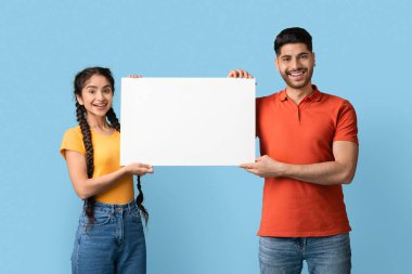 İki neşeli birey, bir erkek ve bir kadın, açık mavi bir arkaplan önünde büyük boş bir tabela tutarak birlikte dururlar. Heyecanlı görünüyorlar ve bir mesaj ya da duyuru paylaşmaya hazırlar..