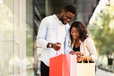Satın Alma Konseptinin tadını çıkarıyorum. Gülümseyen Afrikalı Afrikalı Amerikalı çiftin portresi renkli alışveriş çantalarının içine bakıyorlar, yeni kıyafetleri ve hediyeleri için mutlular, açık havada alışveriş merkezinin yanında duruyorlar.