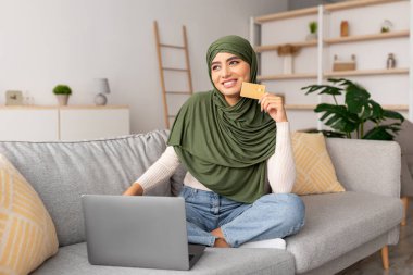 Türbanlı bir Arap kadın kanepesinde bağdaş kurup internetten alışveriş yapmak için dizüstü bilgisayar kullanıyor. Elinde kredi kartı var, rahat bir ortamda evden alışveriş yapıyor..